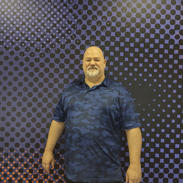 A man with a beard and mustache stands in front of a purple and black dotted background, wearing a blue camo shirt.