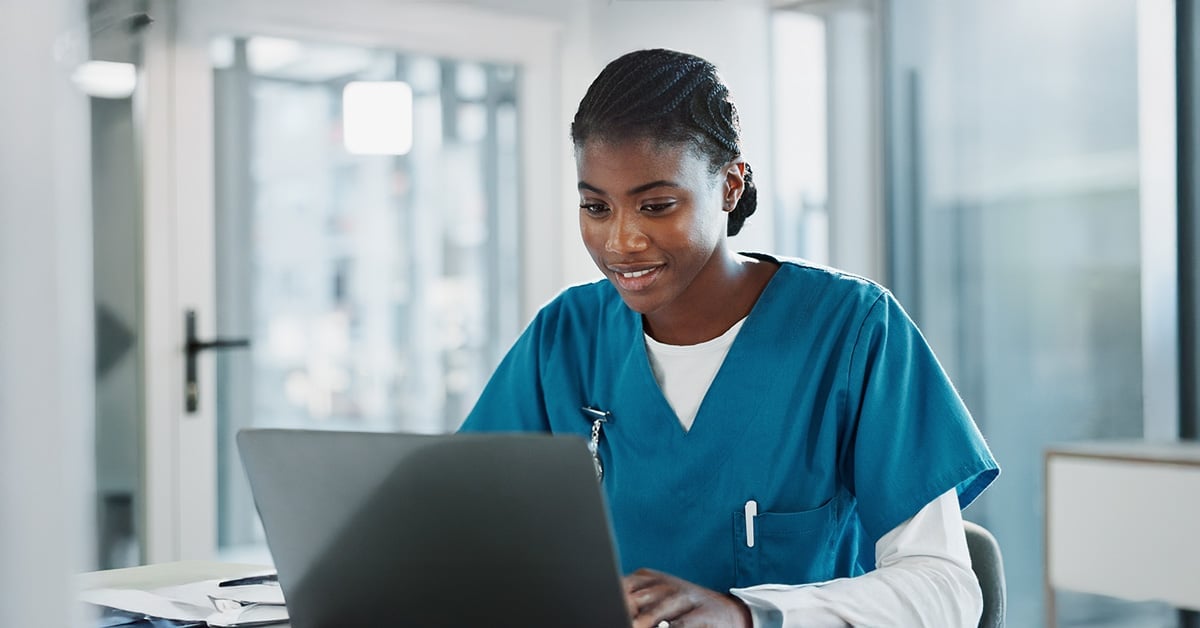 A healthcare professional in blue scrubs is seated at a desk, focused on a laptop. They are smiling and appear to be in a hospital or clinic setting, with windows and medical equipment in the background.