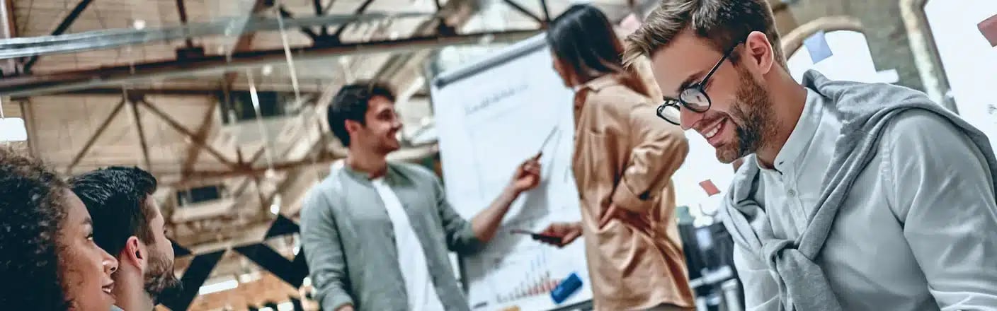 A group of people collaborating in an office, with one person presenting charts on a whiteboard in the background.