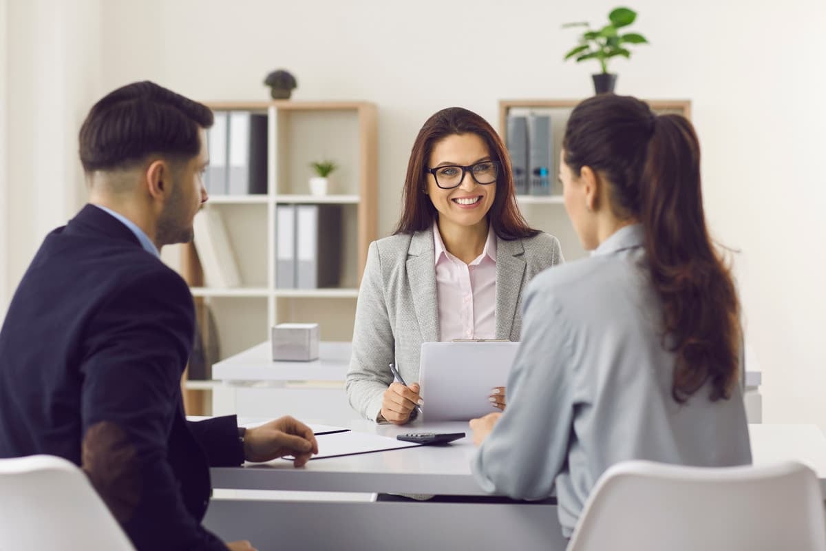 Three people in a business meeting, seated at a table in a bright office. One woman is holding a clipboard and smiling.