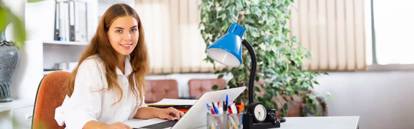 Woman with long brown hair working on a laptop at a desk with a blue lamp, plants, and stationery in the background.