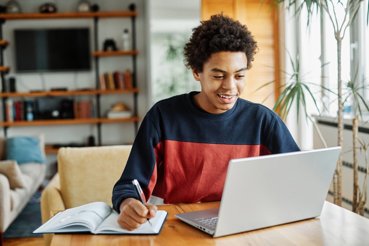 Young person smiling while studying at home, taking notes from a laptop with a pen and open notebook.