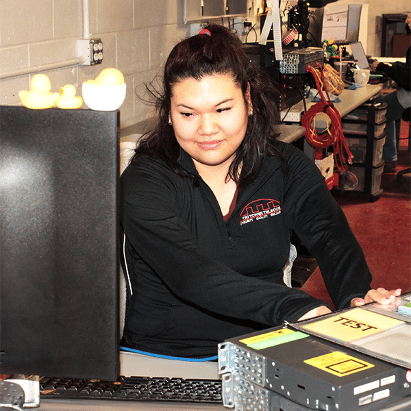 Person seated at a workstation, adjusting equipment beside a monitor with small rubber ducks on top.