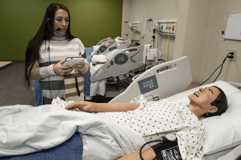 A woman wearing a headset stands beside a hospital bed, observing and holding a tablet, while a medical mannequin in a hospital gown lies in the bed with medical equipment nearby.