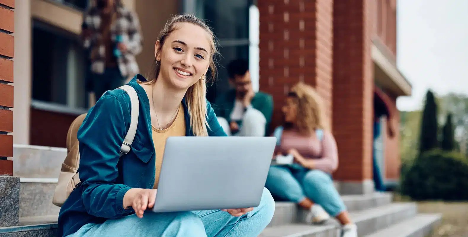 Smiling student sitting on outdoor steps with a laptop, other students in the background near a brick building.