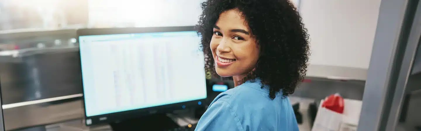 A person in scrubs smiles while sitting in front of a computer screen displaying data, with a blurred background.