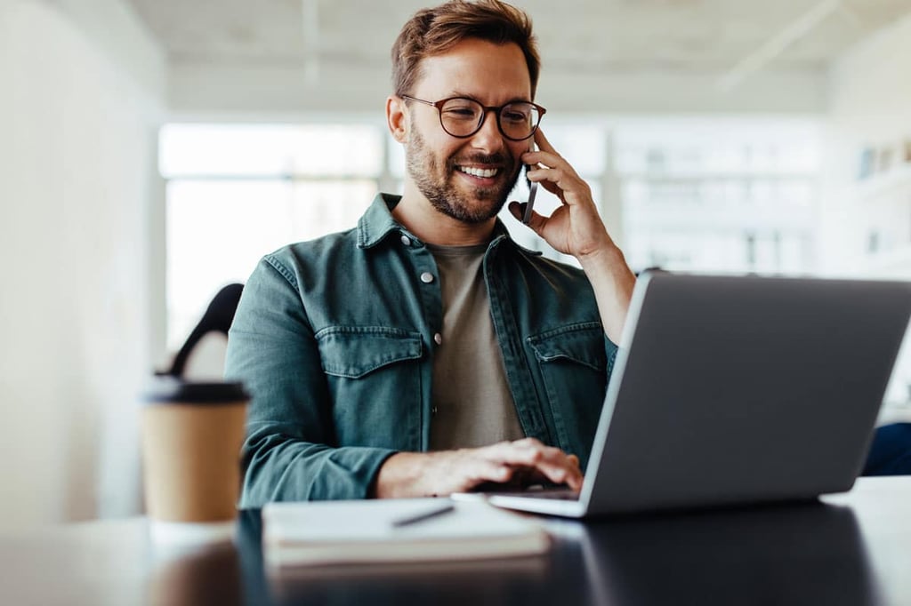 Man wearing glasses smiling while talking on phone and using a laptop at a desk with a coffee cup and notebook.
