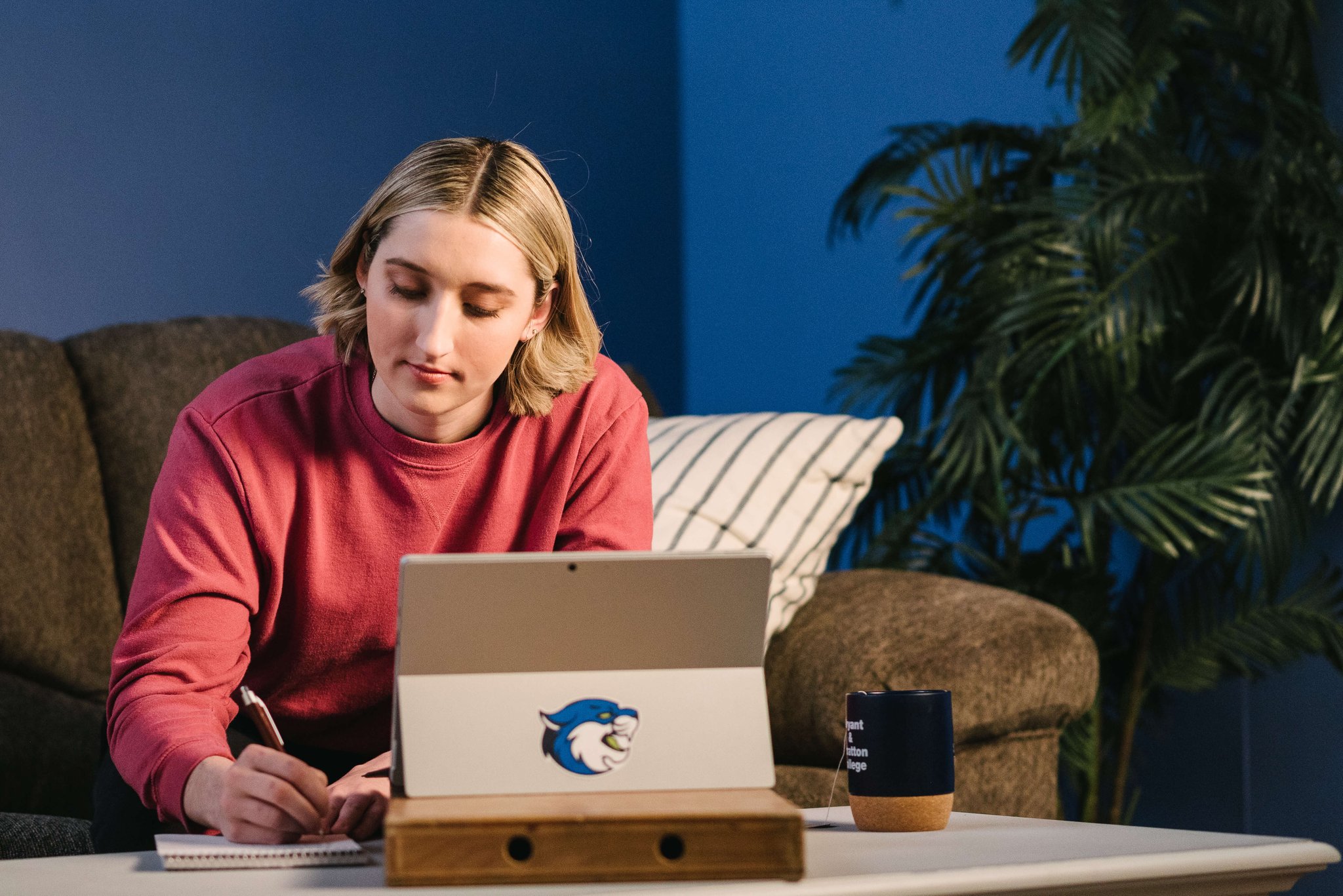 Person in a red sweatshirt writing notes while using a tablet at a desk in a cozy room with a couch and plant.