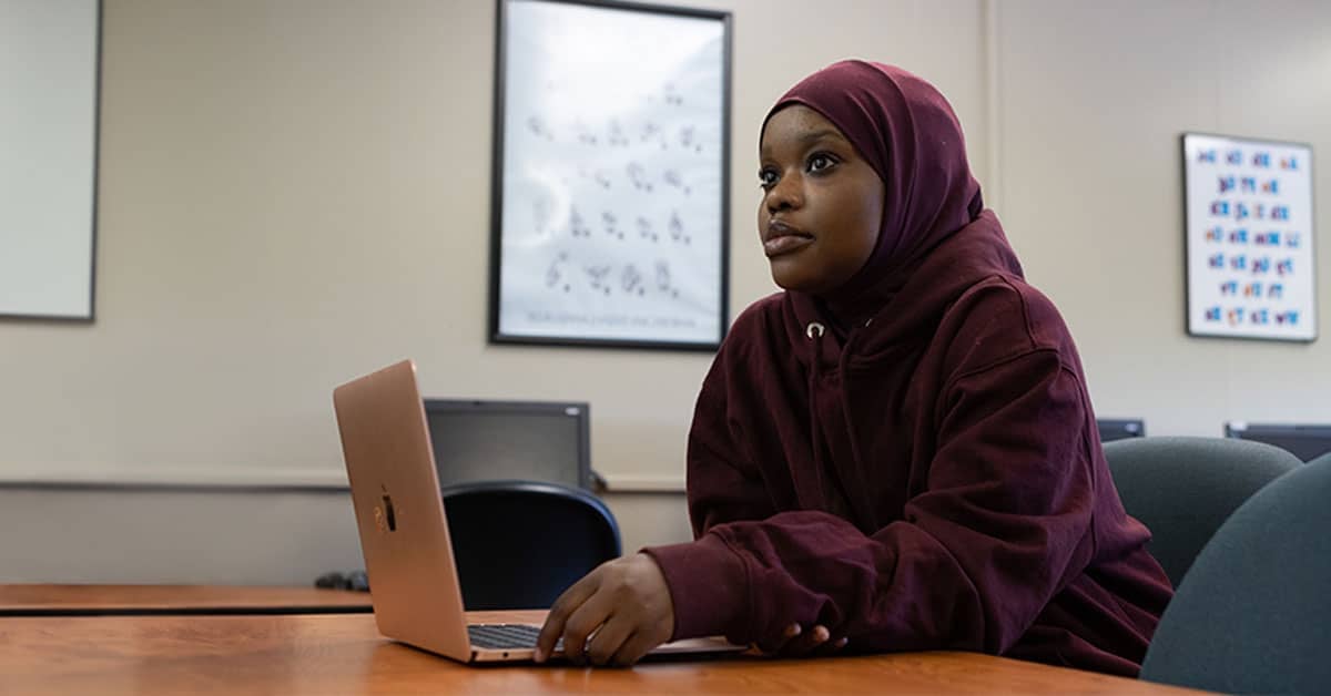 Person in a burgundy hoodie and hijab works on a laptop in a classroom with posters on the wall.