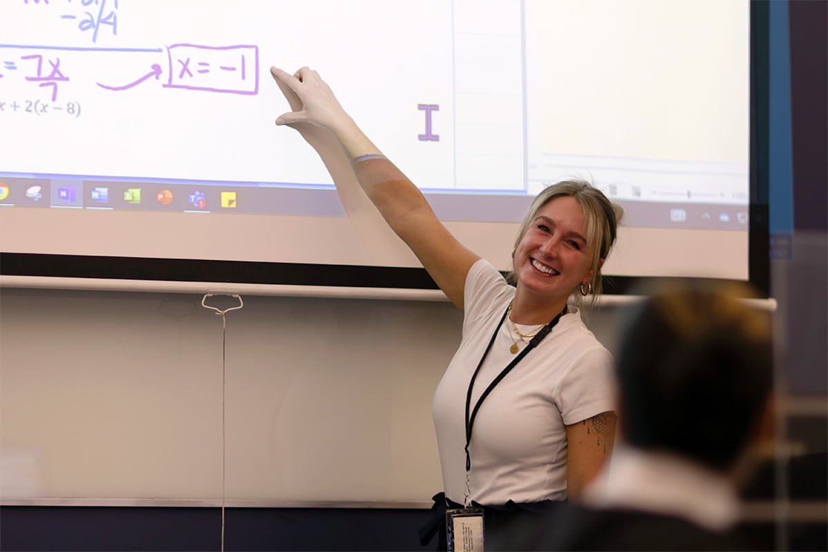 A woman smiling and pointing to a math problem with an equation on a whiteboard during a presentation.