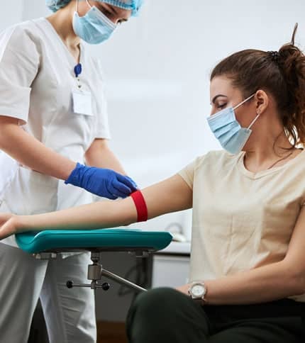 A healthcare professional wearing a mask and gloves prepares to draw blood from a seated woman with a red tourniquet on her arm. Both are wearing masks in a clinical setting.