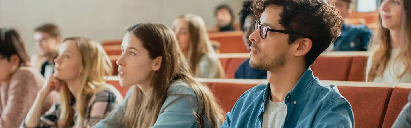 Students attentively listen in a lecture hall, seated in rows, with focus on a young man and woman in the foreground.