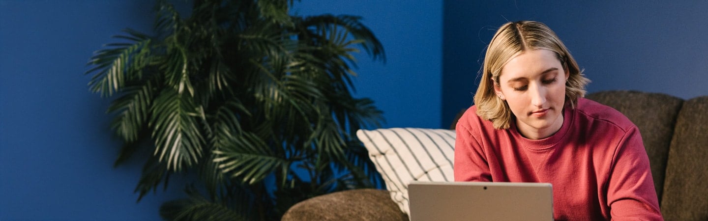 Person in a red sweater sitting on a couch, looking at a laptop, with a large plant and blue wall in the background.