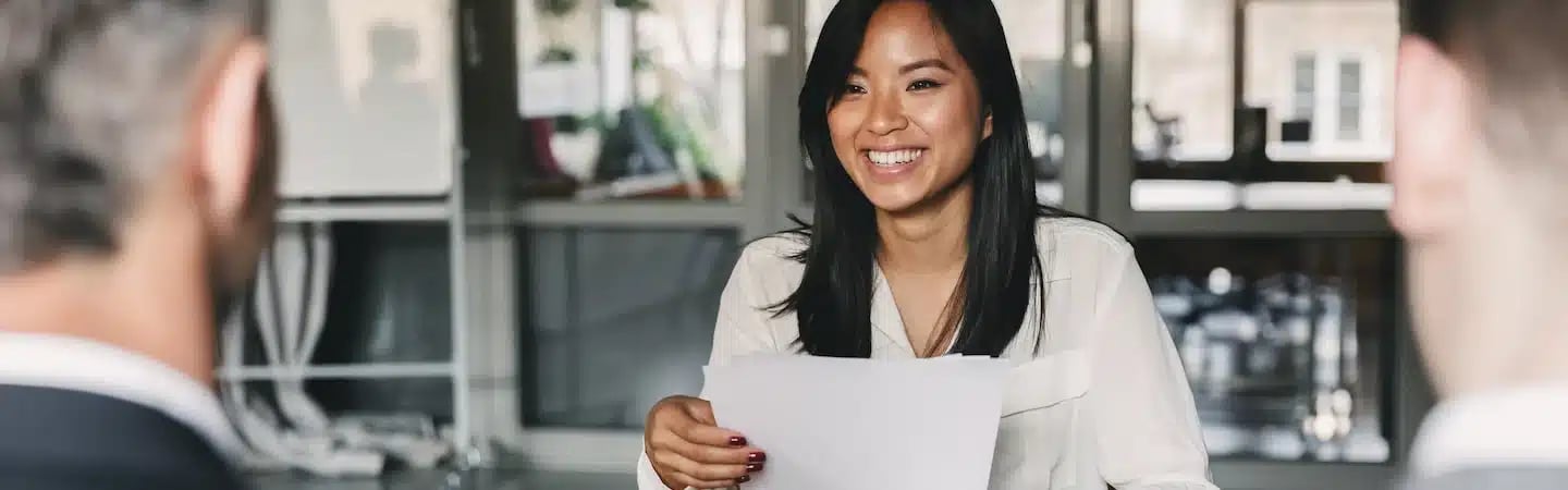 A woman holding papers and smiling while sitting in front of two people during a meeting in an office setting.