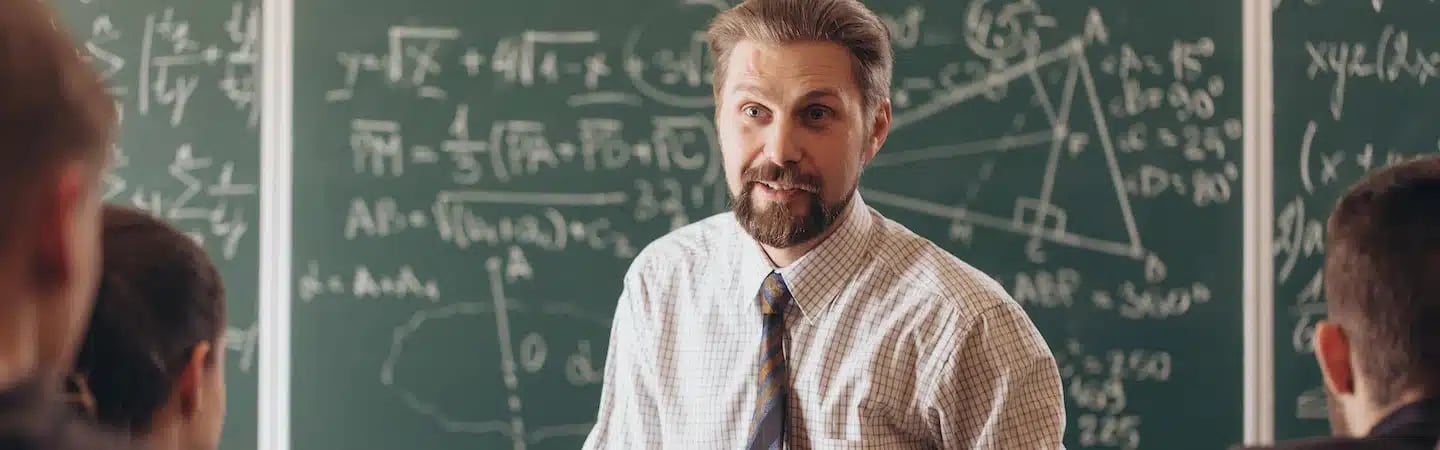 Man in a checked shirt and tie teaching mathematics in front of a chalkboard filled with equations and geometric shapes.