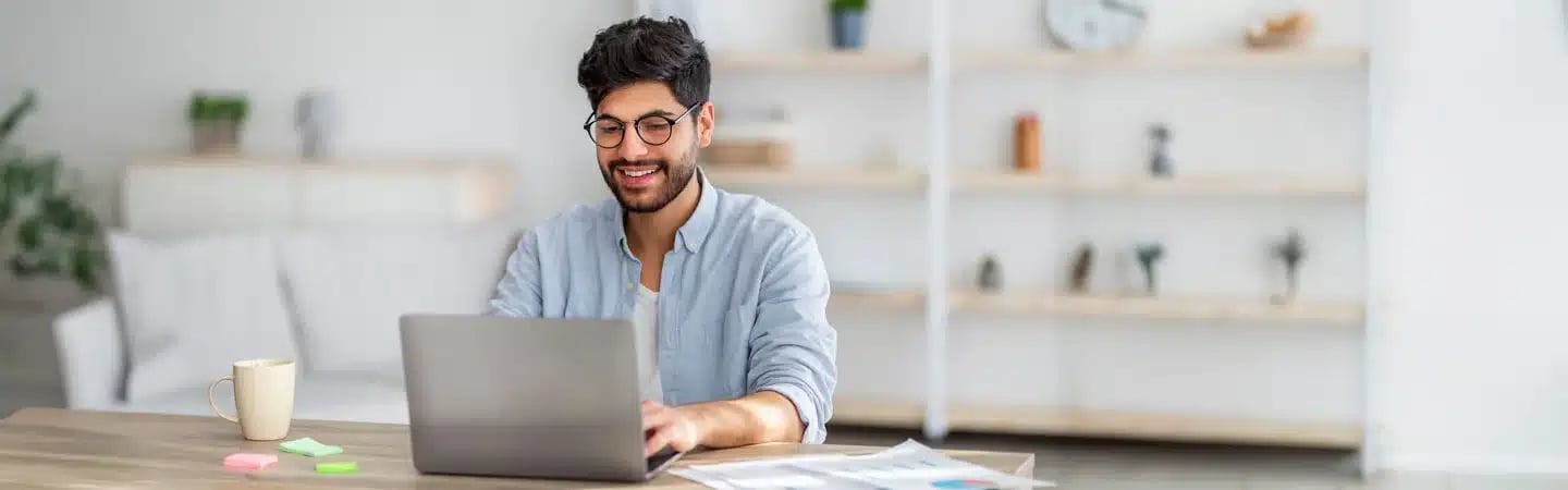 A man wearing glasses and smiling while working on a laptop at a desk with a mug and documents in a well-lit room.