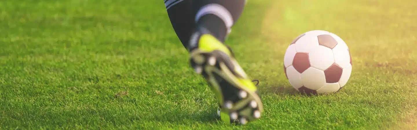 Close-up of a soccer player's legs in motion, kicking a soccer ball on green grass with sunlight in the background.