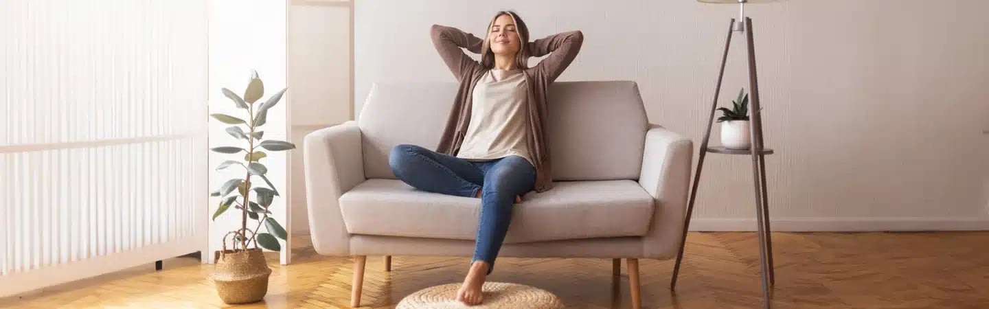 A woman relaxes on a beige sofa in a cozy living room with wooden flooring, plants, and a floor lamp.