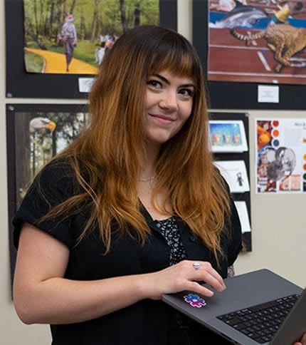 A person with long hair and a black shirt smiles while holding a laptop. Behind them, art and photos are displayed on a wall.
