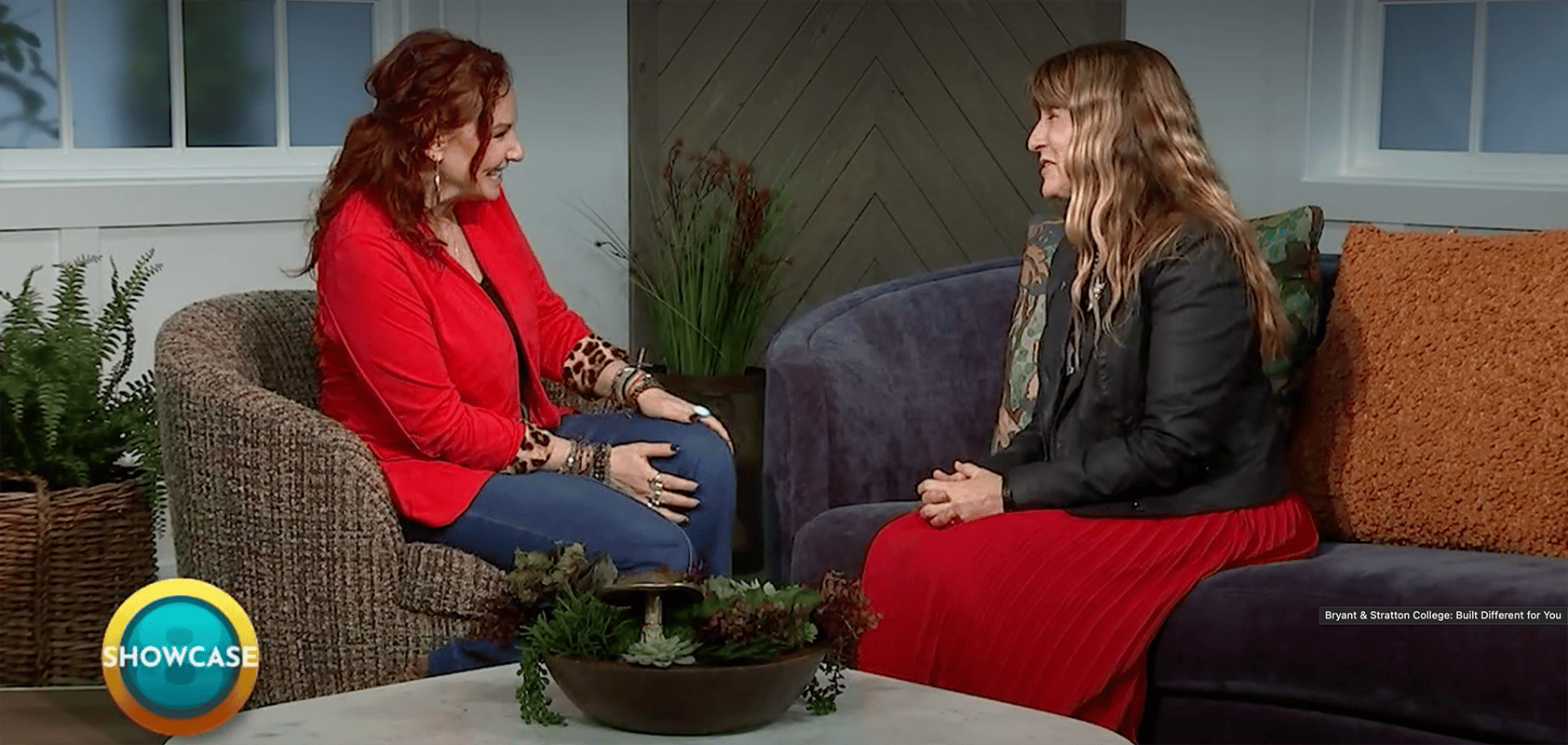 Two women sit facing each other on couches in a cozy studio, engaged in conversation about Rapid Registration at Bryant & Stratton College. One wears a red jacket and jeans, the other a black jacket and red skirt. A "SHOWCASE" logo appears in the bottom left corner.