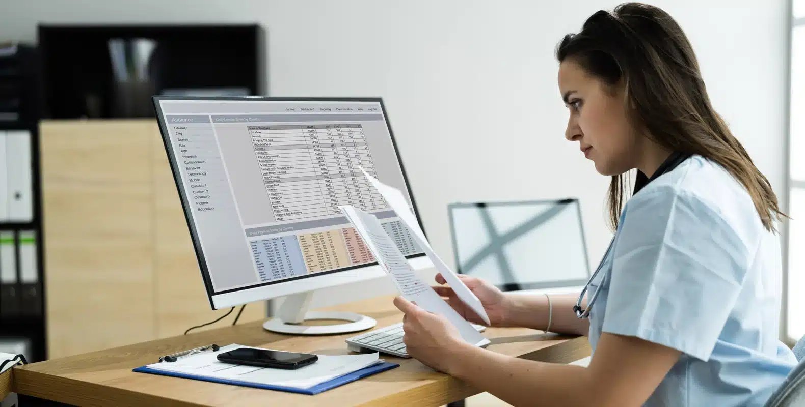 A woman wearing scrubs sits at a desk, reviewing documents in front of a computer displaying a spreadsheet.