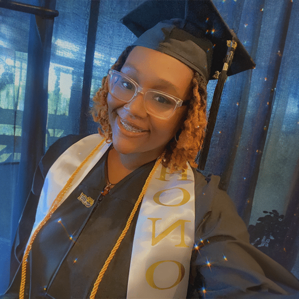 A graduate wearing a cap and gown with a white honor stole smiles in front of a blue curtain.