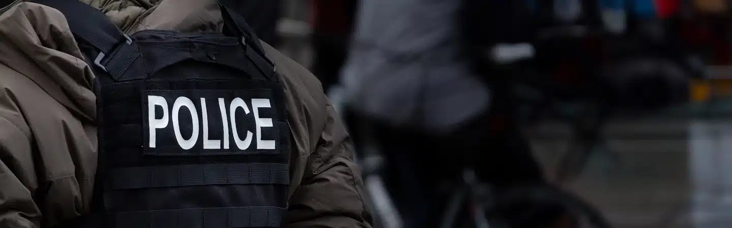 A person dressed in a jacket with a vest labeled "POLICE" stands facing away, with blurred figures in the background.