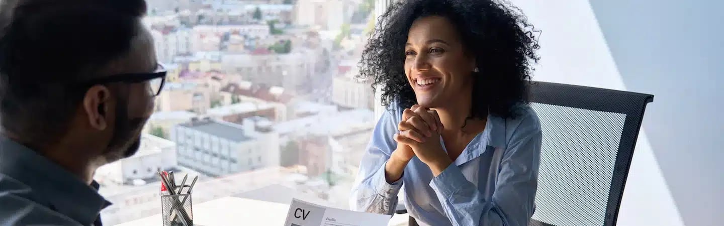 Woman smiling during an office interview, holding a resume. Bright cityscape visible through the large window behind them.