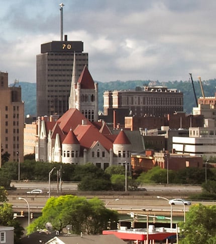 The Syracuse skyline with prominent buildings and lush greenery under a partly cloudy sky.