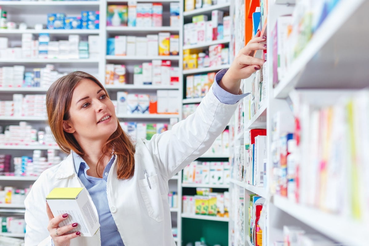 Pharmacist in white coat reaching for medicine on a shelf, holding a medication box in a well-stocked pharmacy.