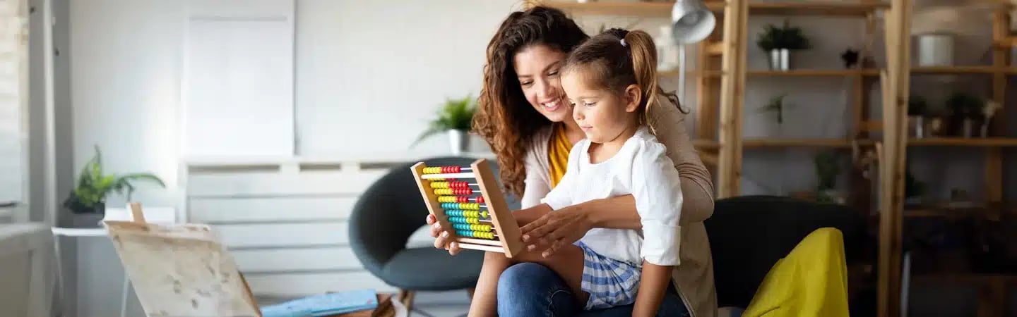 A woman and a young girl sit together, using a colorful abacus in a cozy, well-lit room with plants and shelves.