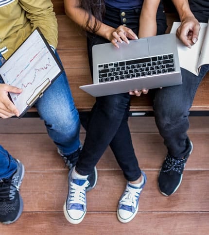 Three people sitting on a wooden bench, one holding a laptop, another holding a notebook, and the third showing a clipboard with a financial chart. Only their lower bodies and hands are visible.