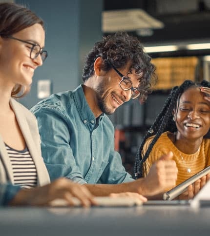 Three people are sitting at a table, smiling and engaged in discussion. One is wearing glasses and a striped shirt, another has curly hair and glasses, and the third has braided hair and is holding a tablet. They appear to be in a cheerful, collaborative setting.