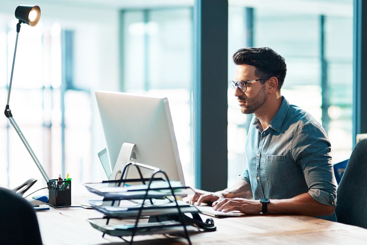 Man working diligently at a computer in a brightly lit office with a desk lamp and organized files.