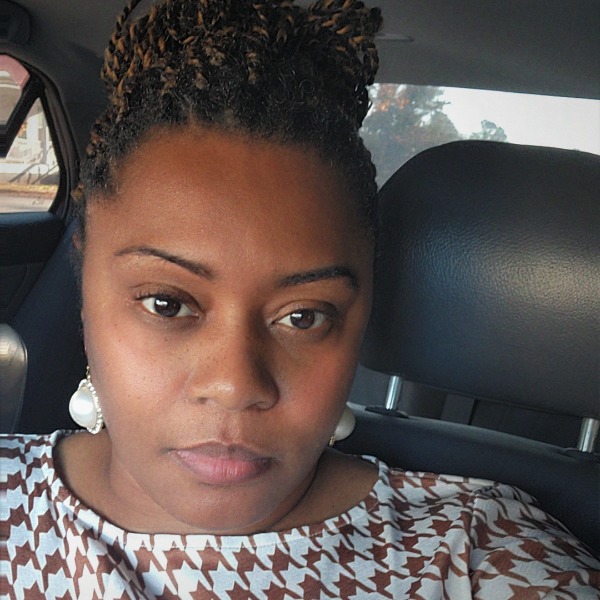 A woman with braided hair sits in a car in Richmond, wearing a patterned top and large pearl earrings, looking directly at the camera with a neutral expression—her confident demeanor hinting at her background in Health Services Administration and recent achievement of an LPN Diploma.