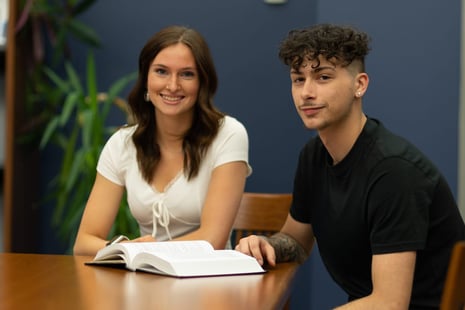 Two people sit at a table, smiling with an open book in front of them. The background features plants and a blue wall.