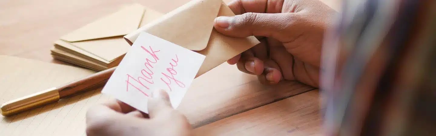 Person holding a thank you note and envelope with more envelopes and a fountain pen on a wooden table.