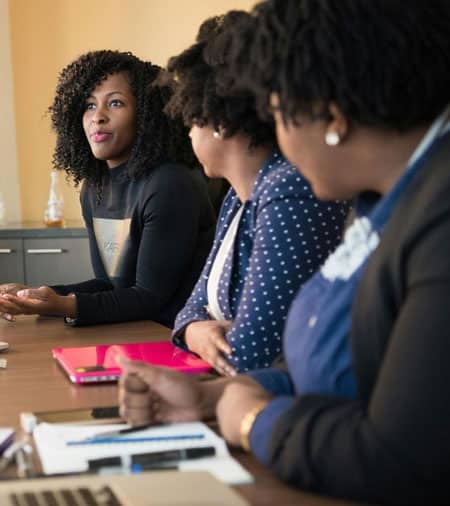 Three women sit at a conference table engaged in discussion. One woman, facing the group, gestures as she speaks, while the others listen attentively. Laptops, papers, and pens are on the table.