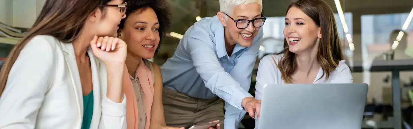 A diverse group of four women smile and collaborate around a laptop at a modern office.