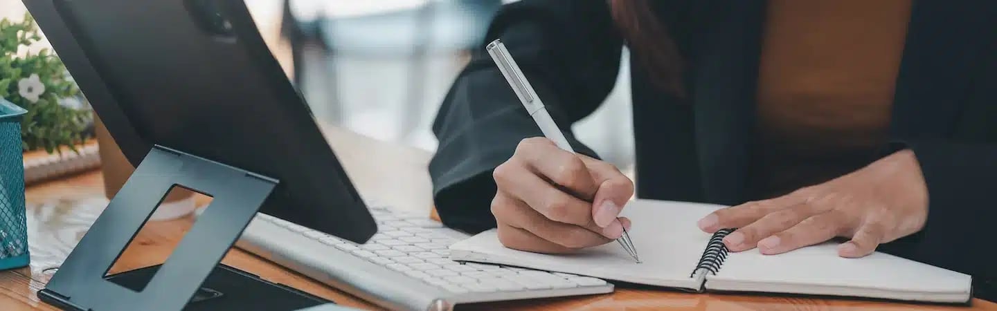 Person writing in a notebook with a silver pen, next to a tablet and keyboard on a wooden desk.