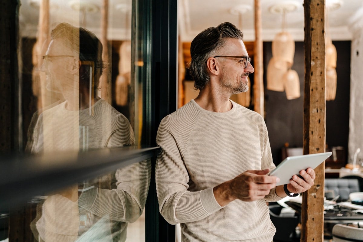 Man in a light sweater holding a tablet, standing by a glass wall, looking to the side with a thoughtful expression.