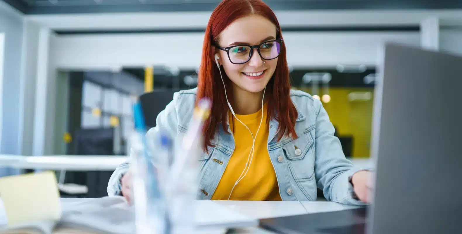 Woman with red hair and glasses smiling while working on a laptop, wearing earphones in a modern office setting.