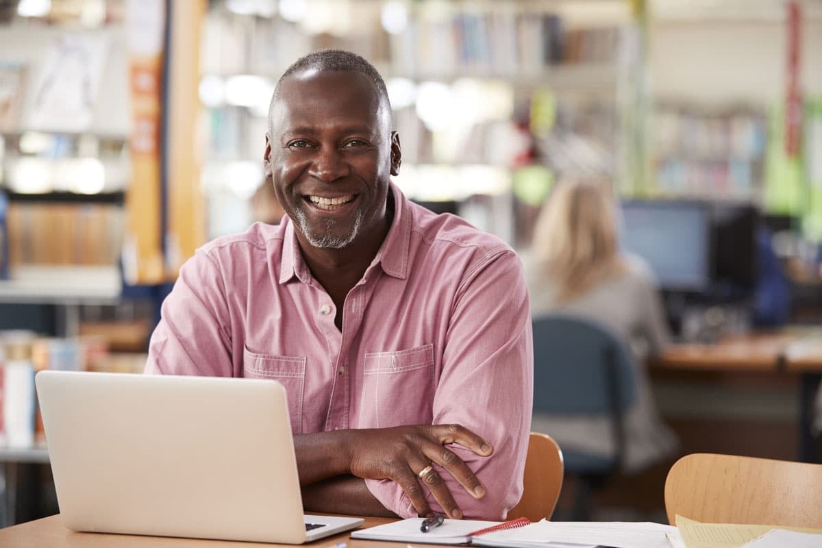 Smiling man with a gray beard sitting at a desk with a laptop in a library or office setting.