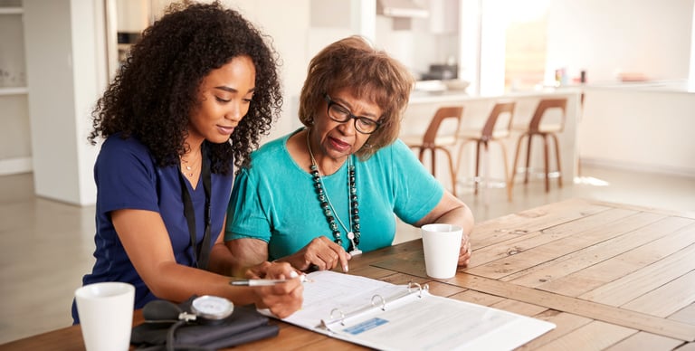 A younger woman and an older woman sit at a table with coffee mugs, reviewing documents in a binder together in a bright, modern kitchen.