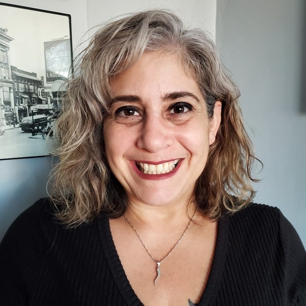 Smiling woman with wavy gray hair and a black sweater poses in front of a vintage photograph background.