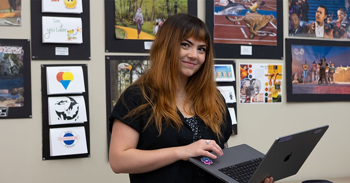 A woman holding a laptop stands in front of a wall display filled with various art pieces and photographs.