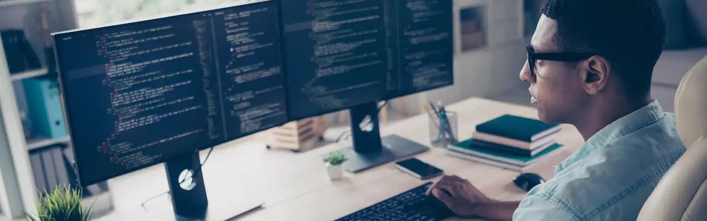 A person is working on a computer with dual monitors displaying code, seated at a desk with books and a potted plant.