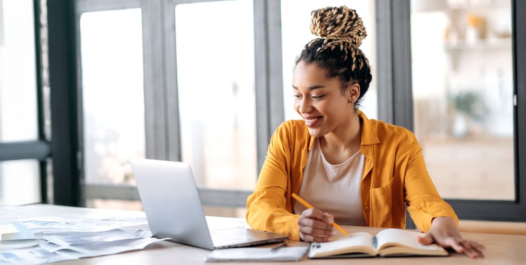 A woman sits at a desk using a laptop, smiling as she writes in a notebook with a pencil. She wears a yellow shirt and has her hair in a bun. Sunlight streams through large windows in the background.
