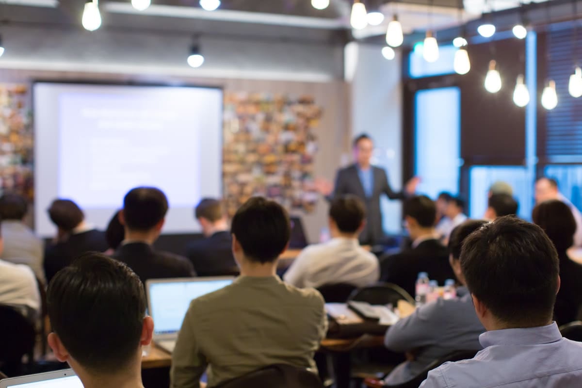 People seated in a conference room, watching a presentation on a screen, with lights hanging from the ceiling.