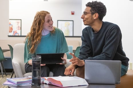 Two people are sitting and smiling at each other, one holding a tablet and the other pointing, with books and laptops nearby.